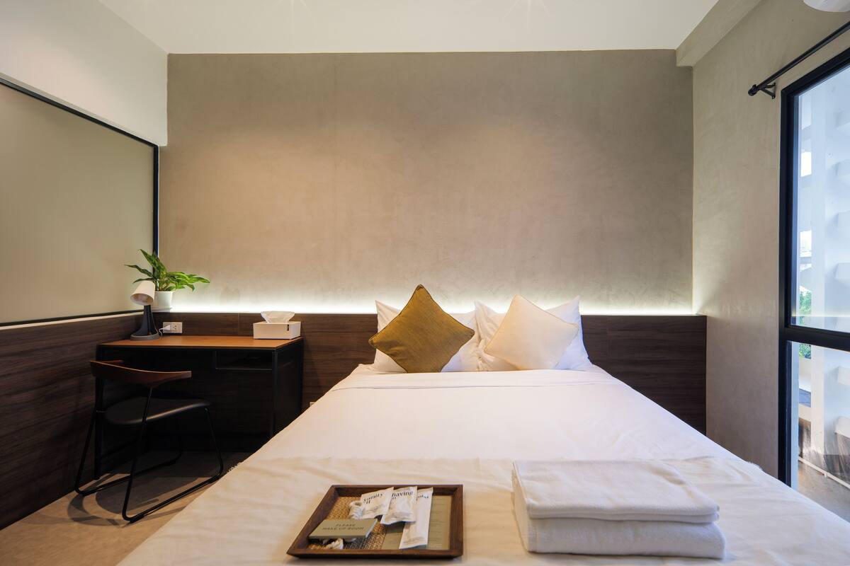 A modern hotel room with a large bed, a desk with a chair, a potted plant, and a window providing a view of the outdoors.
