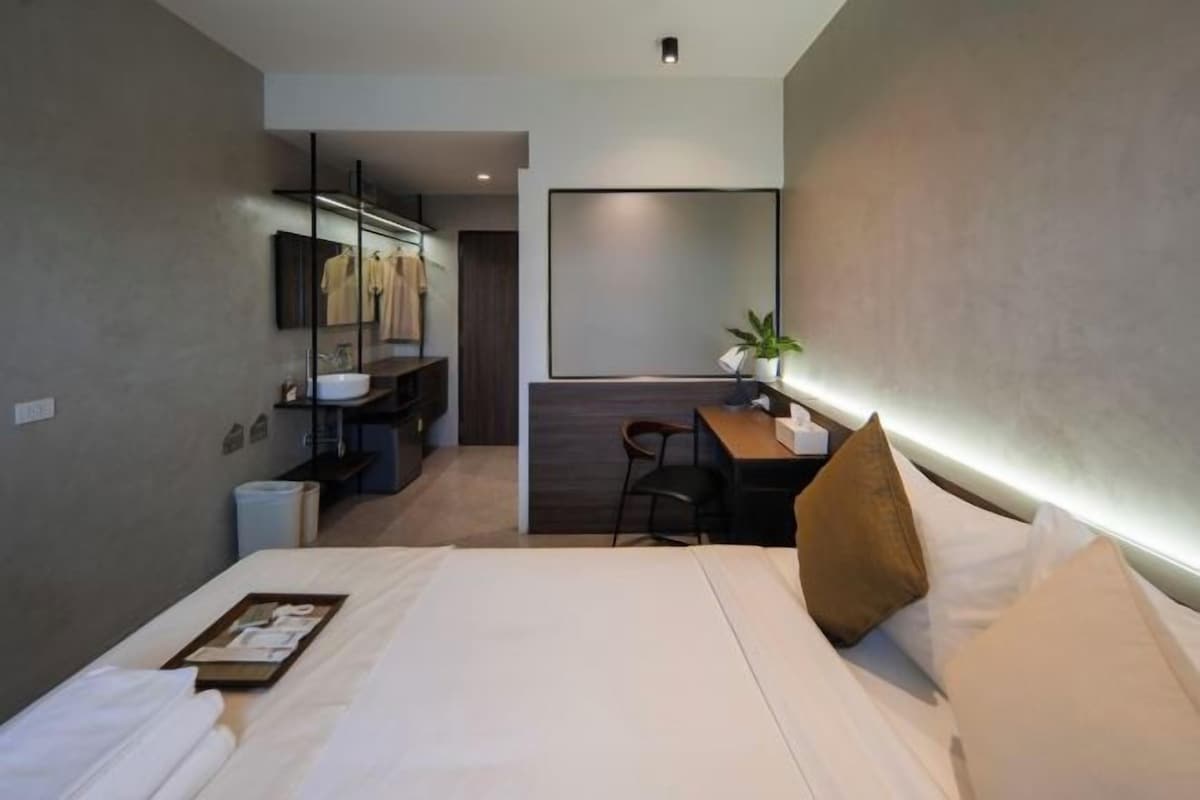 A modern hotel room with a large bed, a desk with a chair, a small table, a mirror, and a plant.