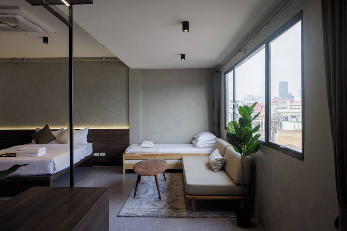 A modern hotel room with a bed, a sofa, a small table, and a large window offering a city view.