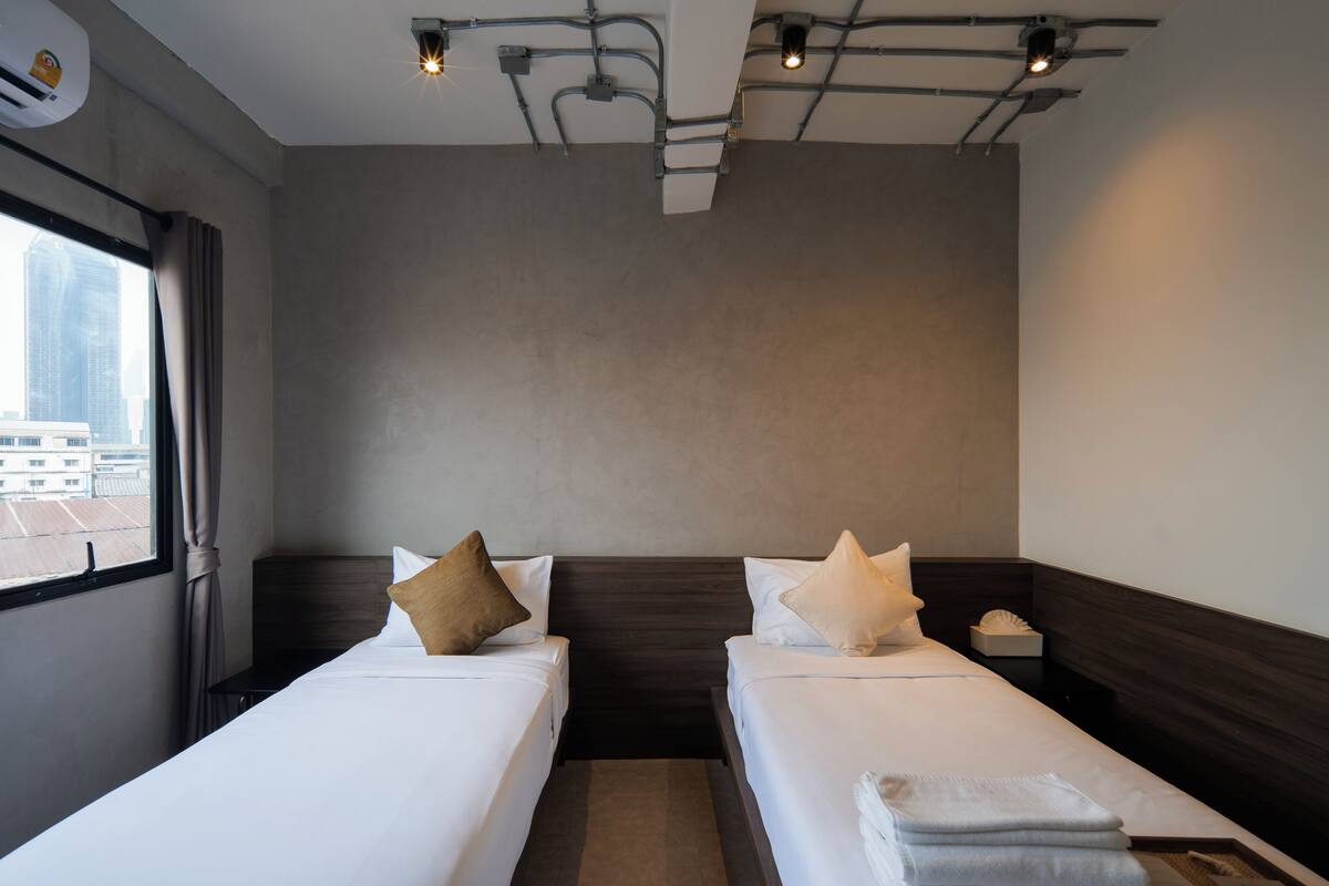 A hotel room with two beds, a window with curtains, a ceiling with exposed pipes, and a city view.