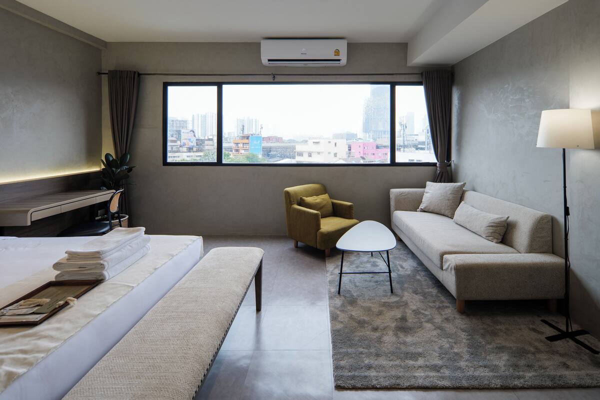 A hotel room with a bed, a sofa, a chair, a small table, and a large window offering a city view.