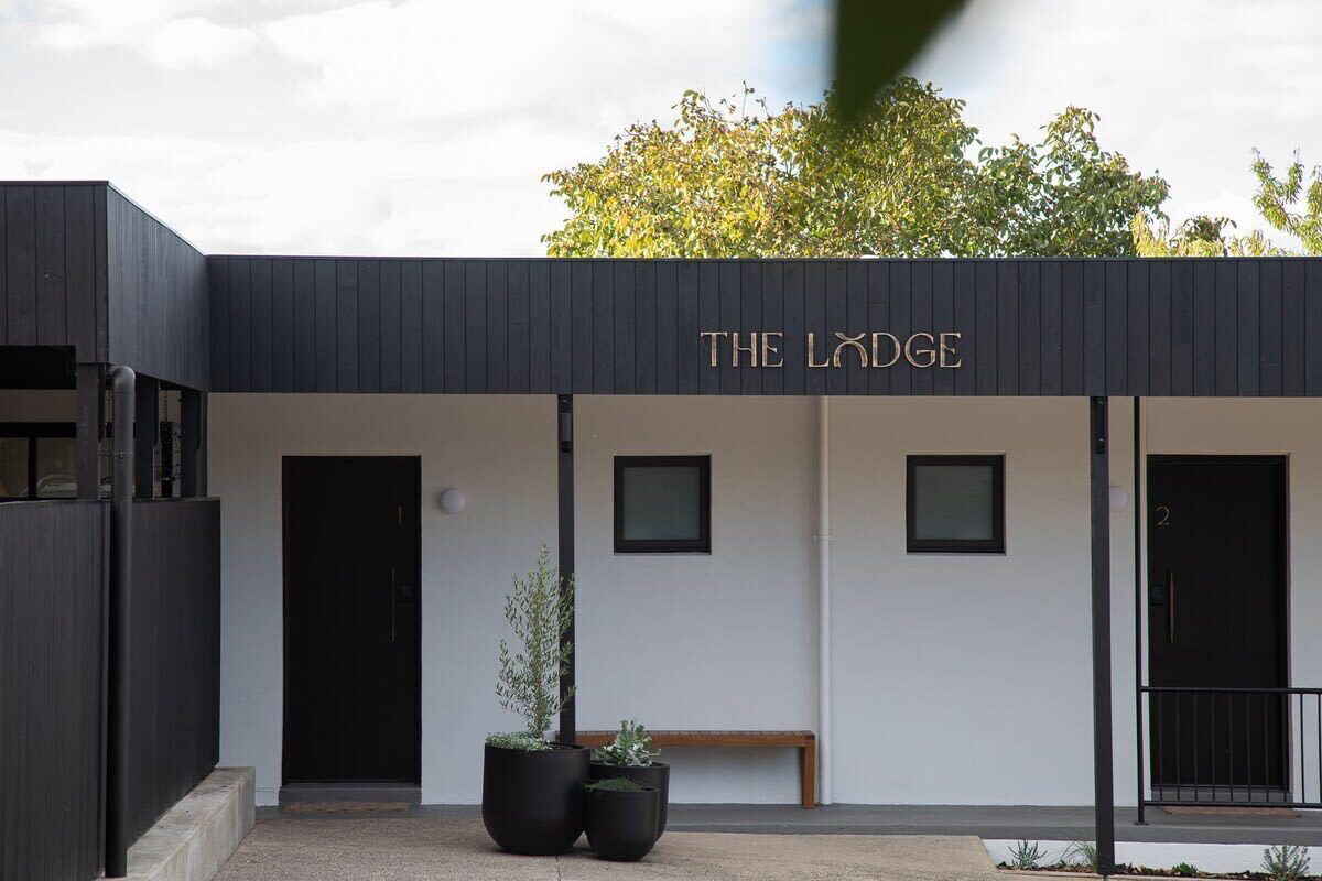 Een gebouw genaamd "The Lodge" met een minimalistisch ontwerp, met twee grote zwarte deuren, een bankje en twee potplanten.