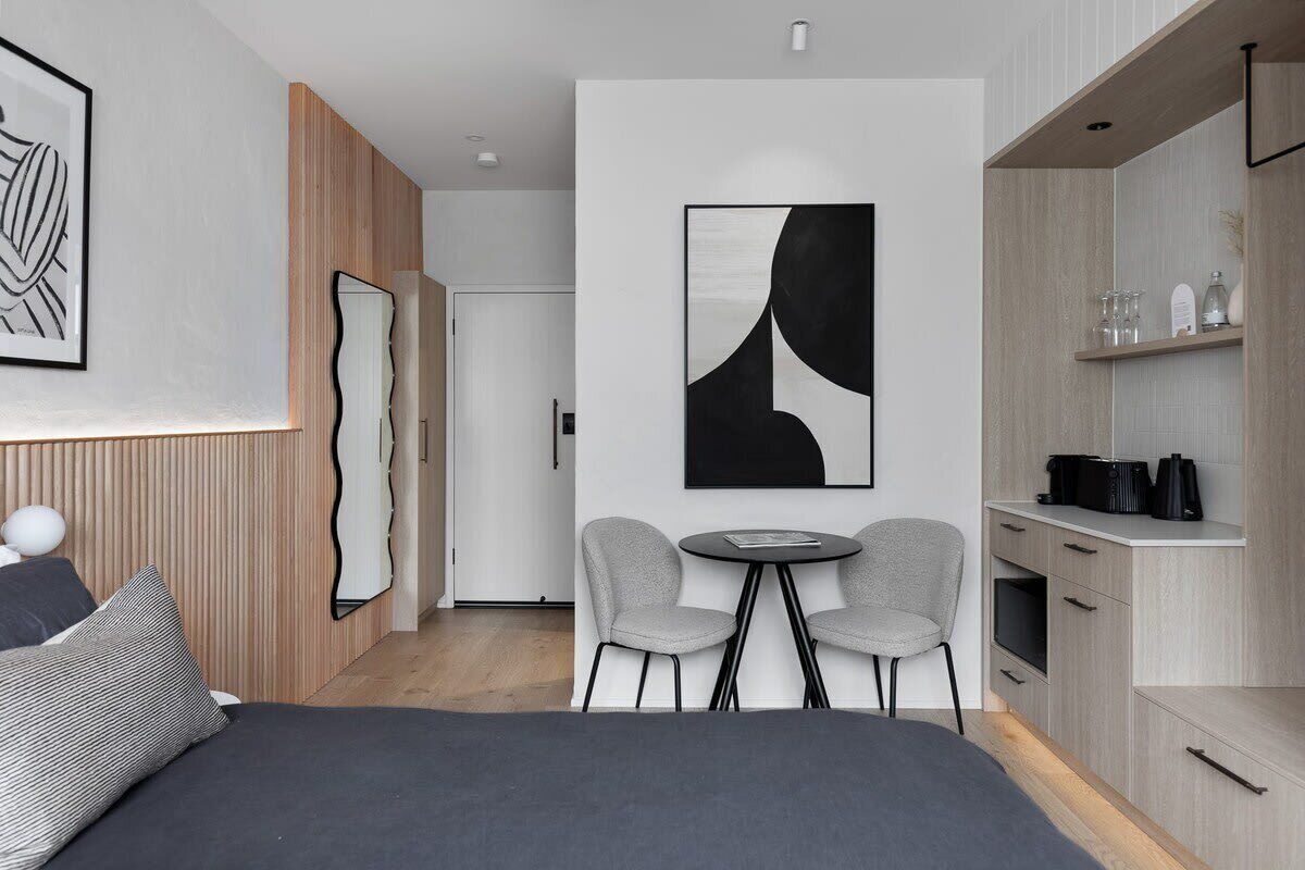 A modern hotel room with a bed, a small dining table with chairs, a kitchen area, and artwork on the walls.