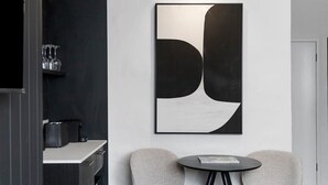 Una habitación moderna con diseño minimalista, que presenta un cuadro abstracto grande, una mesa redonda pequeña, dos sillas y una cafetera sobre una encimera.