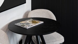 Una mesa redonda negra con una revista encima, ubicada junto a una silla tapizada de color gris, frente a una pared oscura.