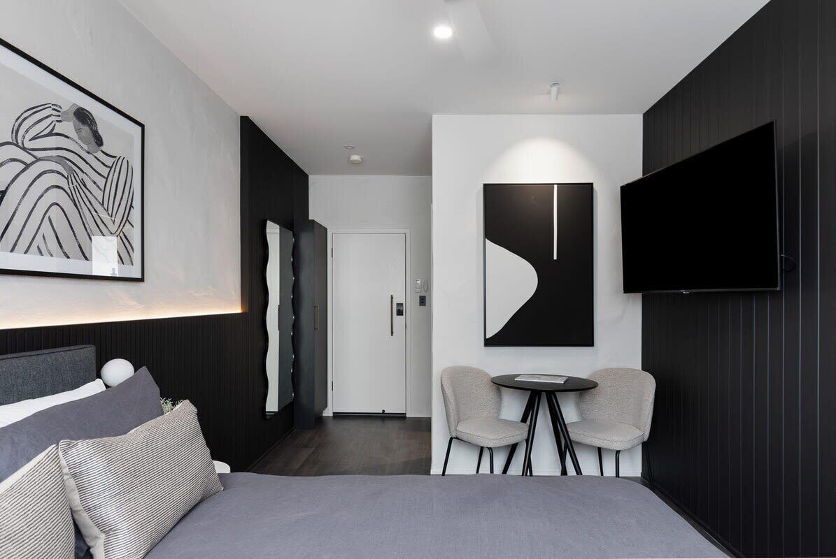 Moderni hotellihuone, jossa on sänky, televisio, pieni pöytä ja kaksi tuolia sekä ovi, joka johtaa toiseen huoneeseen.