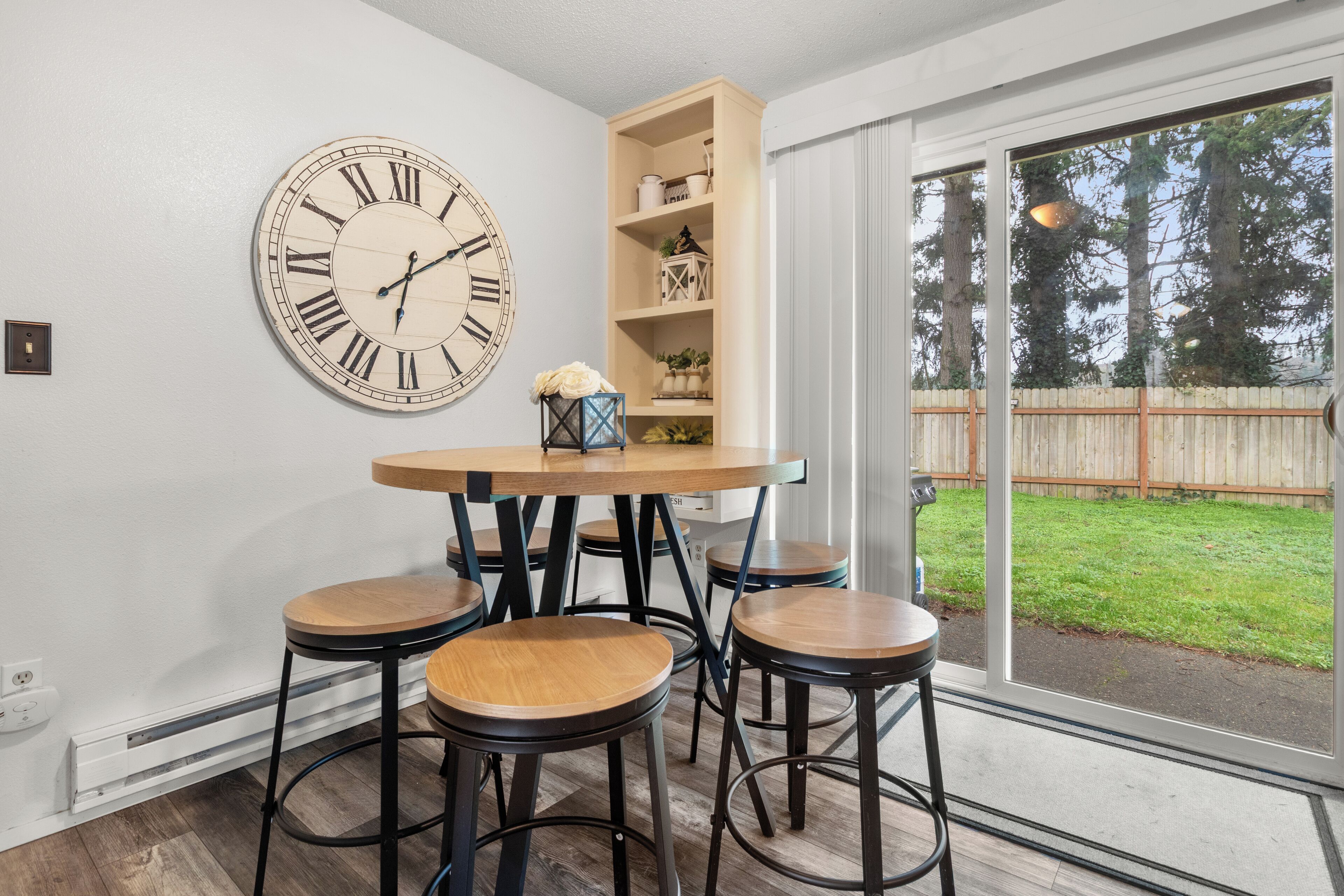 A dining area with a round table, four stools, a large wall clock, and a wooden bookshelf.