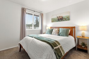 A bedroom with a bed, a window with curtains, a bedside table with a lamp, and a painting on the wall.