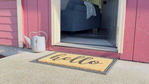 A red house with a blue couch inside, a white kettle outside, and a 'hello' mat on the front door. - Cozy 3BR modern farmhouse in Rochester with stone fireplace, fenced yard, and easy I-5 access. (Rochester)