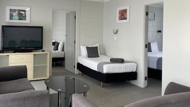 A modern hotel room with a flat-screen TV, two beds, a sofa, and a small table. - Kensington Motel (Whangarei)