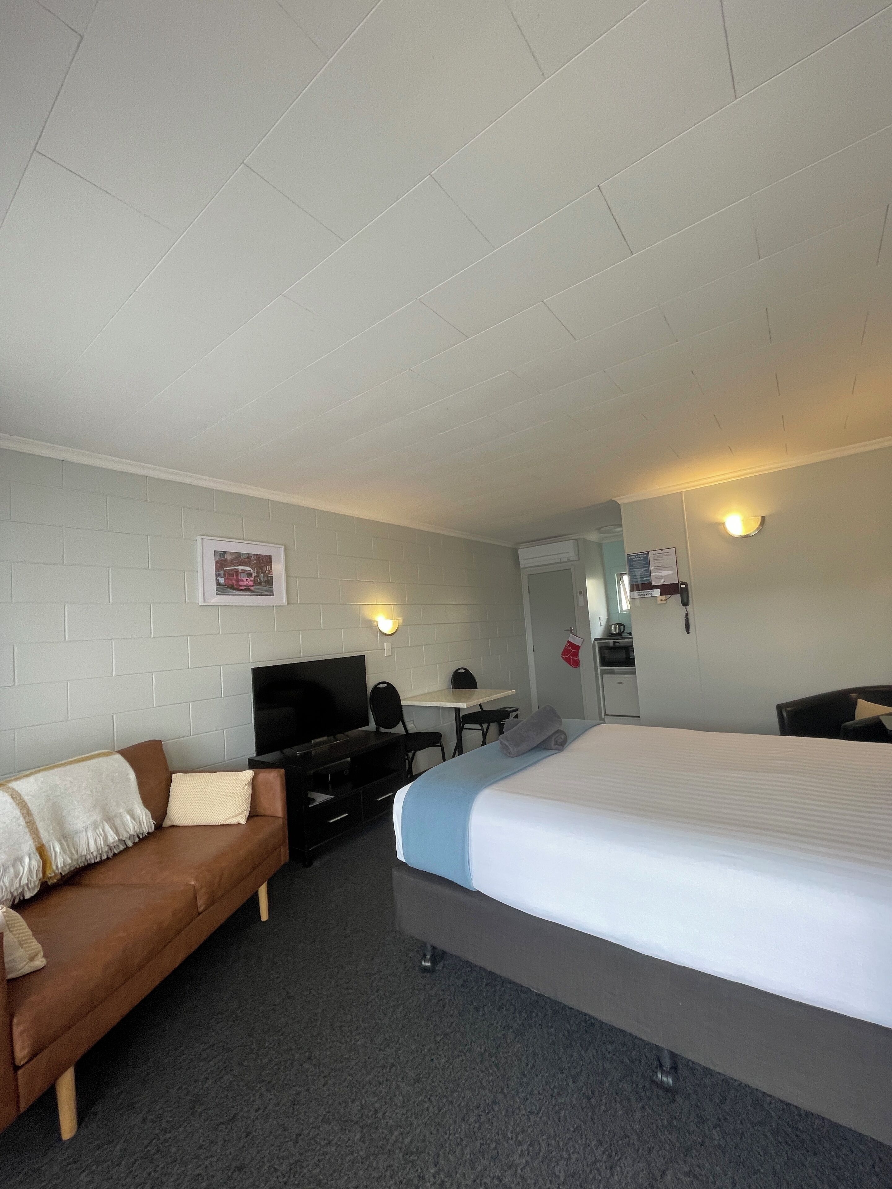 A hotel room with a bed, a sofa, a desk with a chair, a television, and a wall-mounted light fixture.