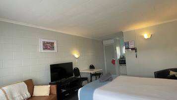 A hotel room with a bed, a sofa, a desk with a chair, a television, and a wall-mounted light fixture.