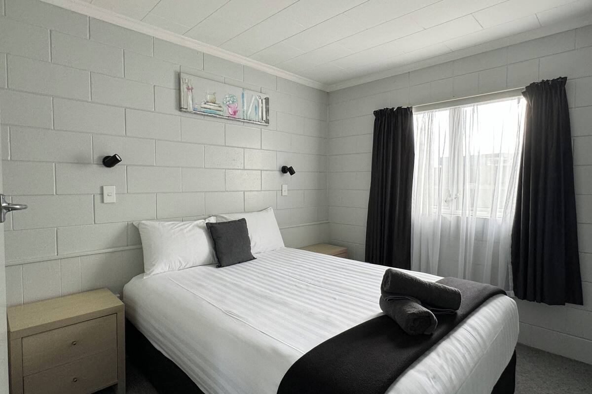 Una habitación de hotel con una cama grande, una mesita de noche, un estante montado en la pared con adornos y una ventana con cortinas.