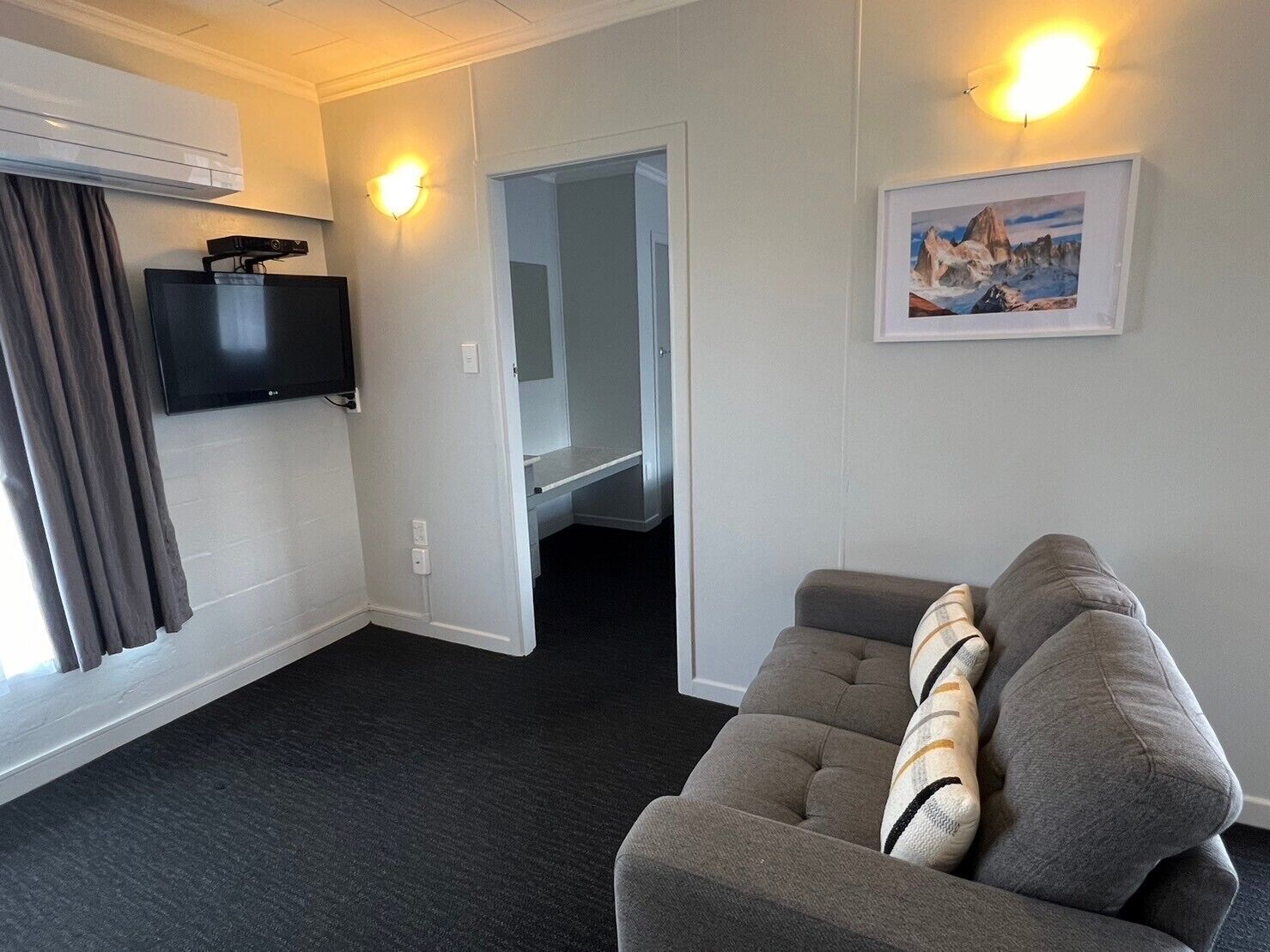 A hotel room with a grey sofa, a flat-screen TV mounted on the wall, a painting on the wall, and a door leading to a hallway.