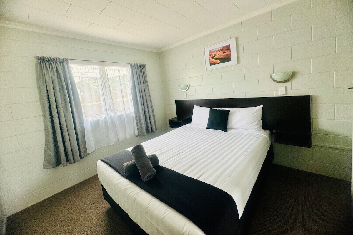 Una habitación de hotel con cama, ventana con cortinas, un cuadro en la pared y una mesita de noche.