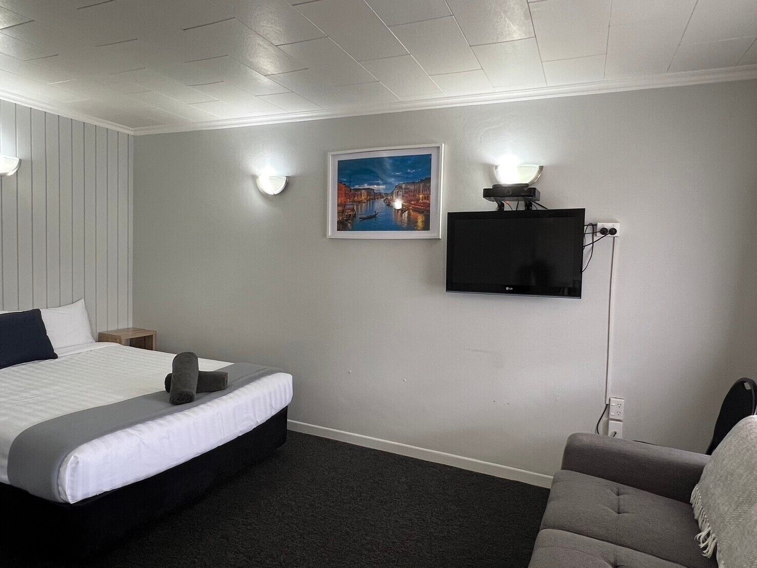Een hotelkamer met een bed, een grijze bank, een flatscreen televisie aan de muur en een ingelijst schilderij boven de televisie.