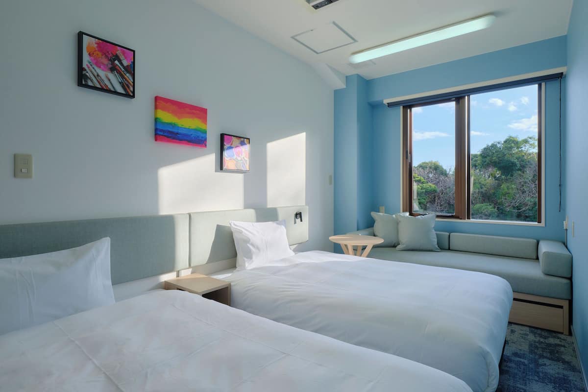 A hotel room with two beds, a small table, a sofa, and a large window with a view of trees.