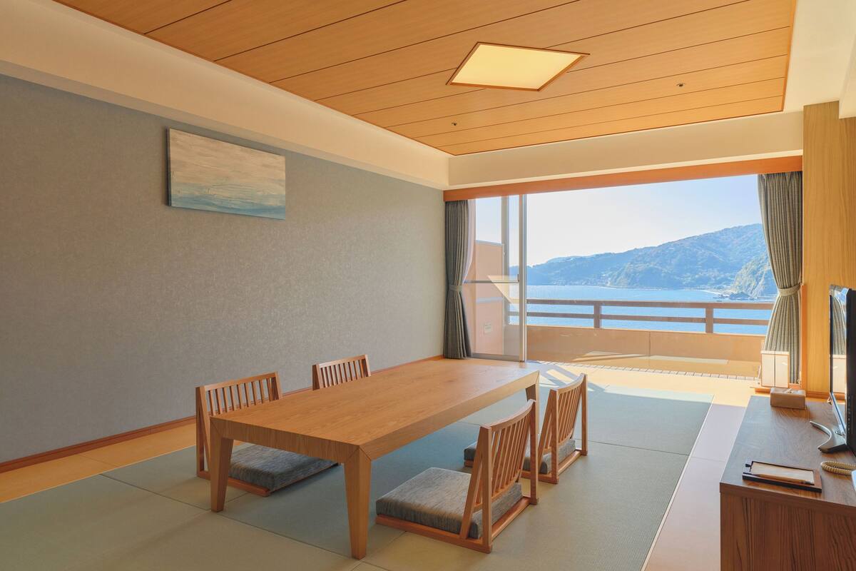 A room with a wooden dining table, four chairs, a television, and a view of a mountainous landscape through large windows.