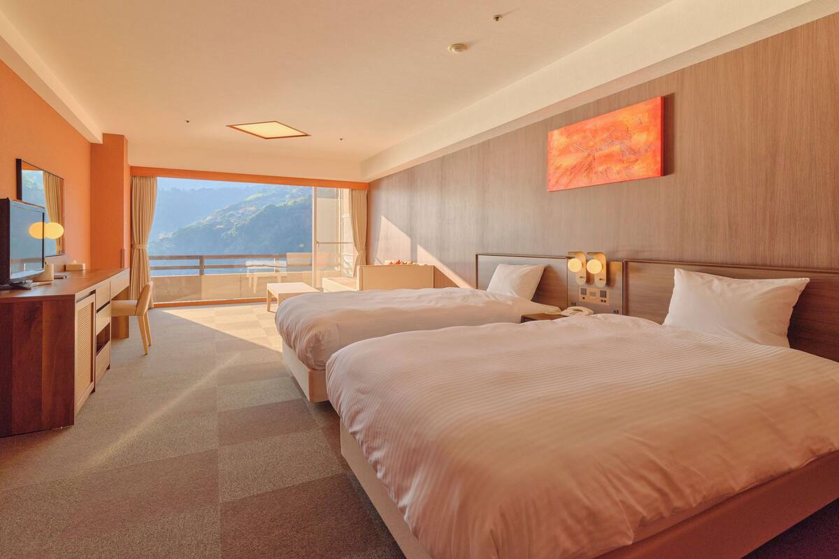 A hotel room with two beds, a desk, a chair, a TV, and a large window with a mountain view.