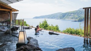 A infinity pool with a view of the sea and hills, a wooden deck, and a stone pathway.