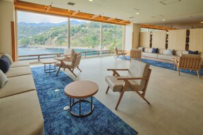 Amplia habitación con un gran ventanal que ofrece vistas a un lago y colinas, amueblada con sofás, sillas y una pequeña mesa.