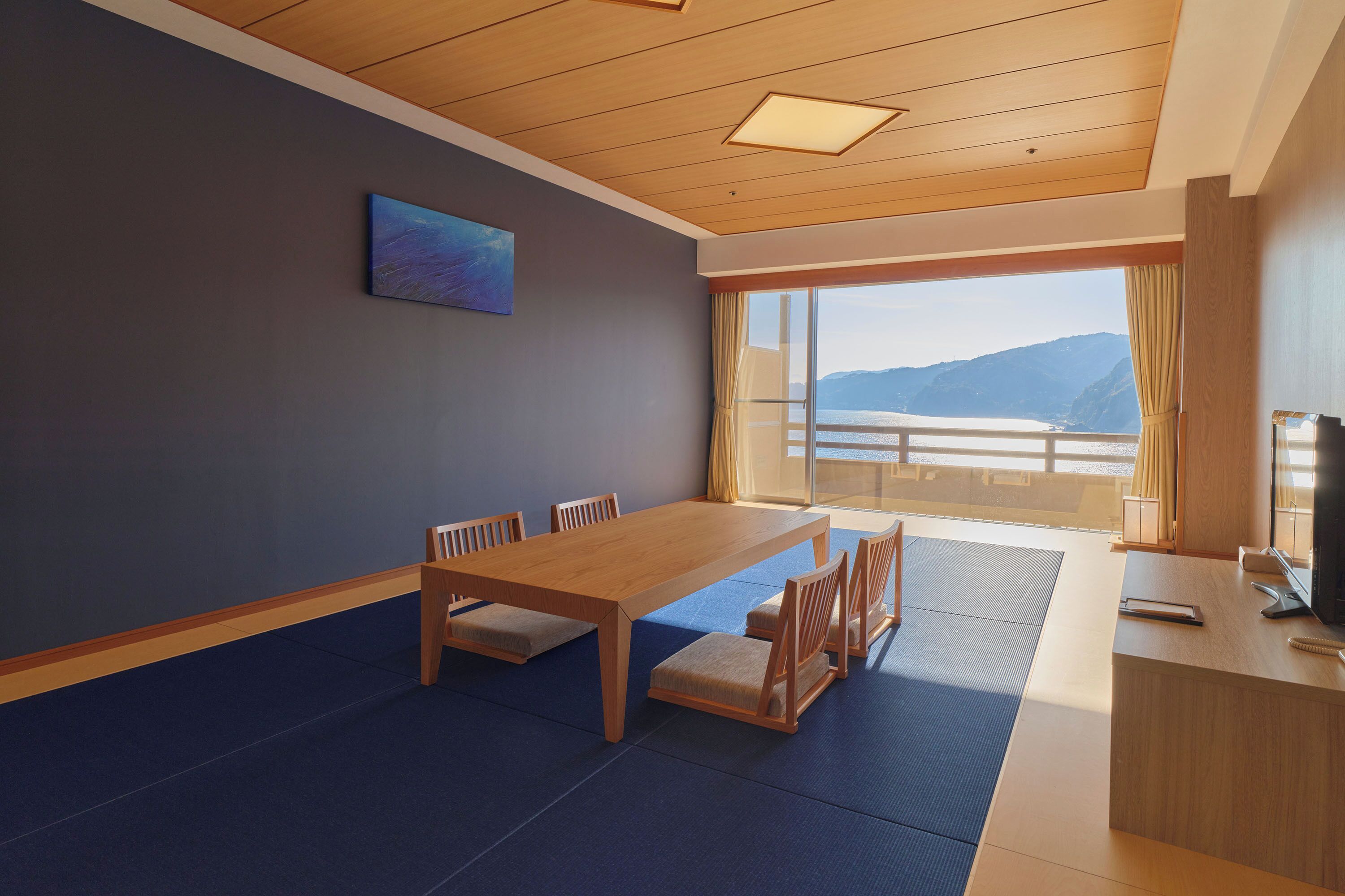 A room with a wooden dining table, chairs, a small desk, a television, and a large window with a view of mountains.
