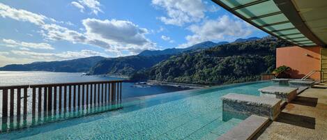 Ein klarer, blauer Pool mit einer Holzterrasse und Stufen, die zu einer Terrasse mit Blick auf ein Gewässer und Berge führen.