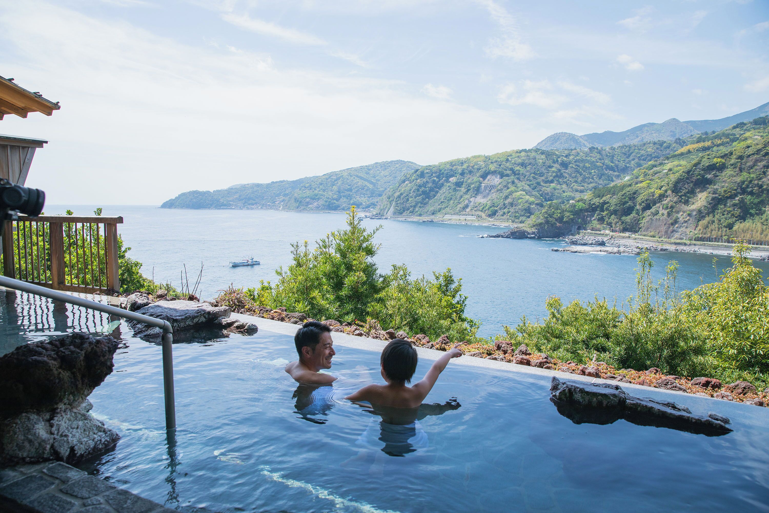 A man and a child in an infinity pool with a view of the sea and mountains.