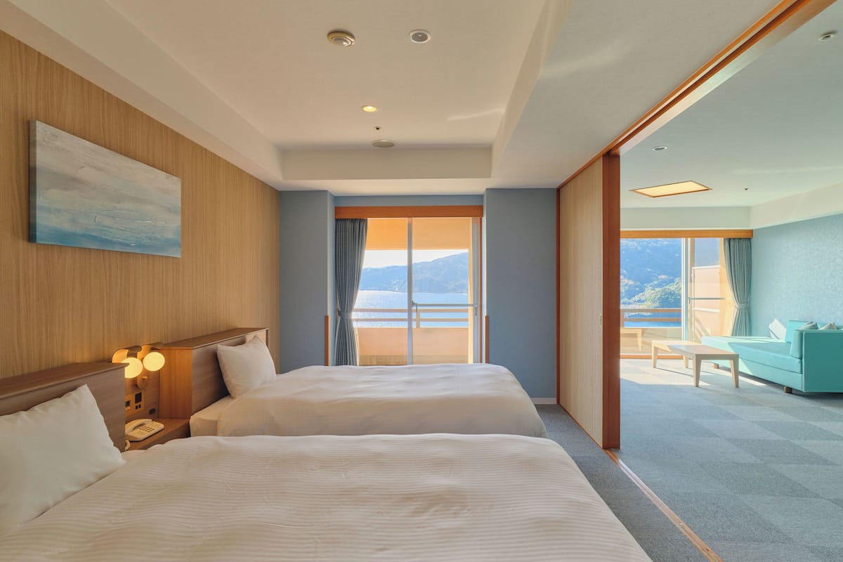 A hotel room with two beds, a nightstand with a phone, a painting on the wall, and a balcony with a view of the mountains.