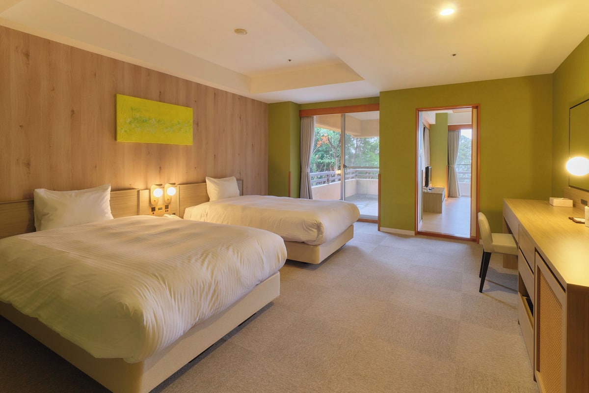 A hotel room with two beds, a desk, a chair, and a balcony with a view.