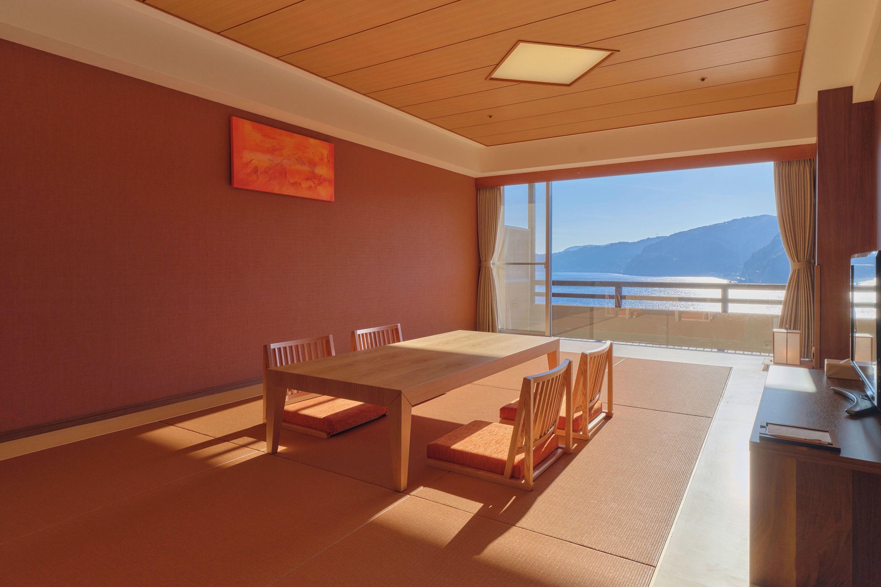 A room with a wooden table and chairs, a painting on the wall, and a large window with a view of mountains and water.