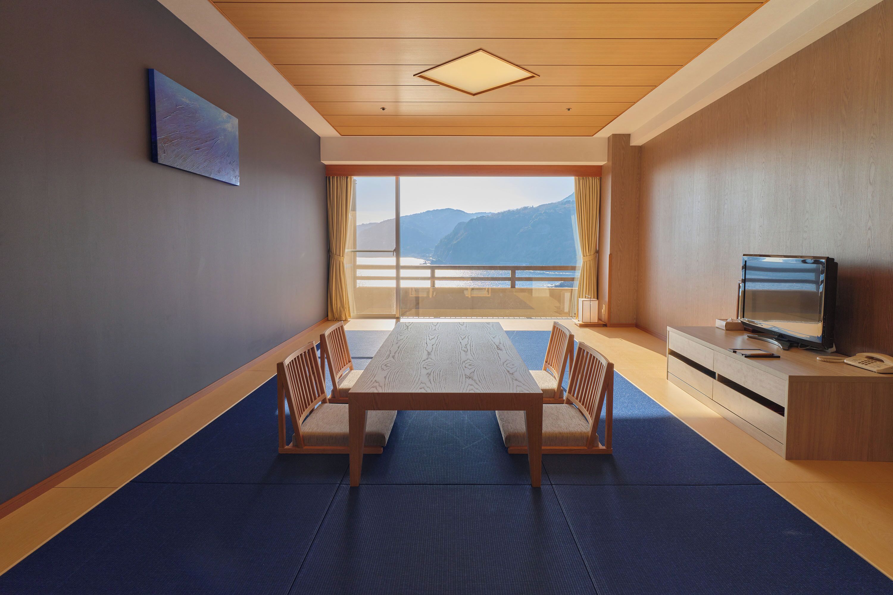 A room with a wooden table, chairs, a TV, and a large window with a view of mountains.
