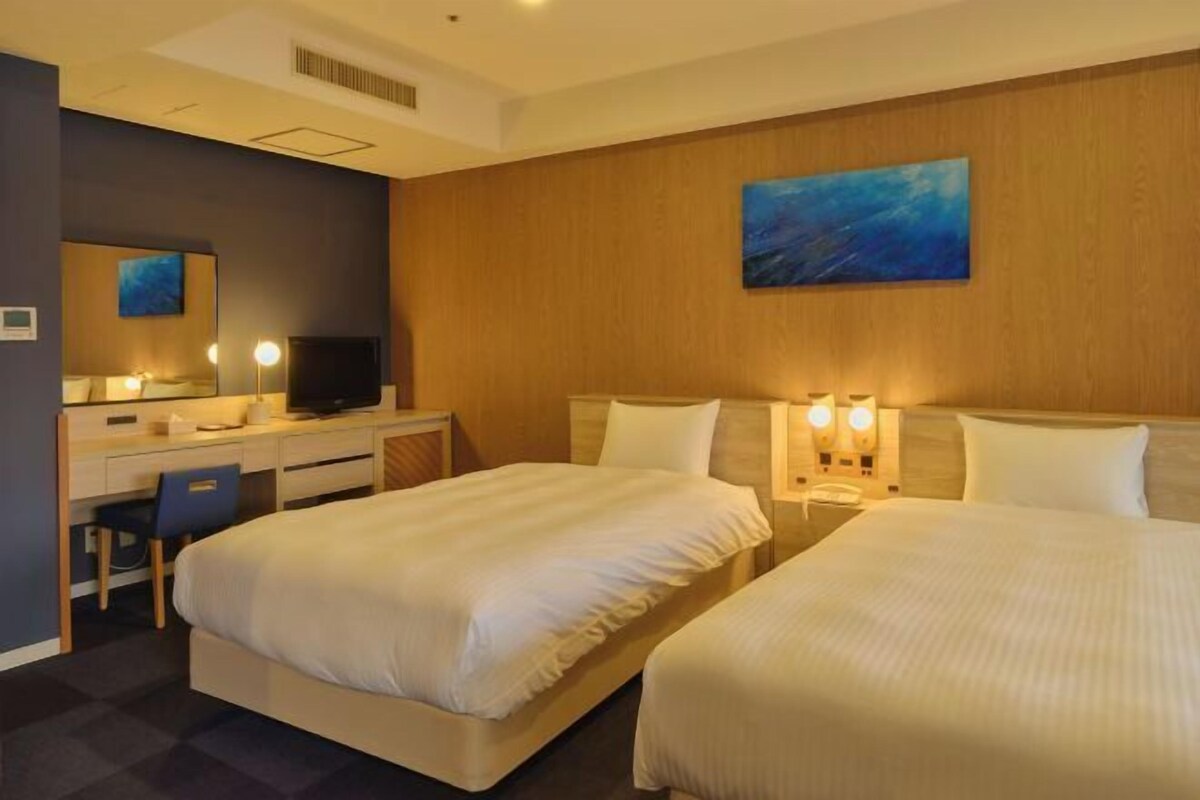 A hotel room with two beds, a desk, a chair, a television, and artwork on the walls.