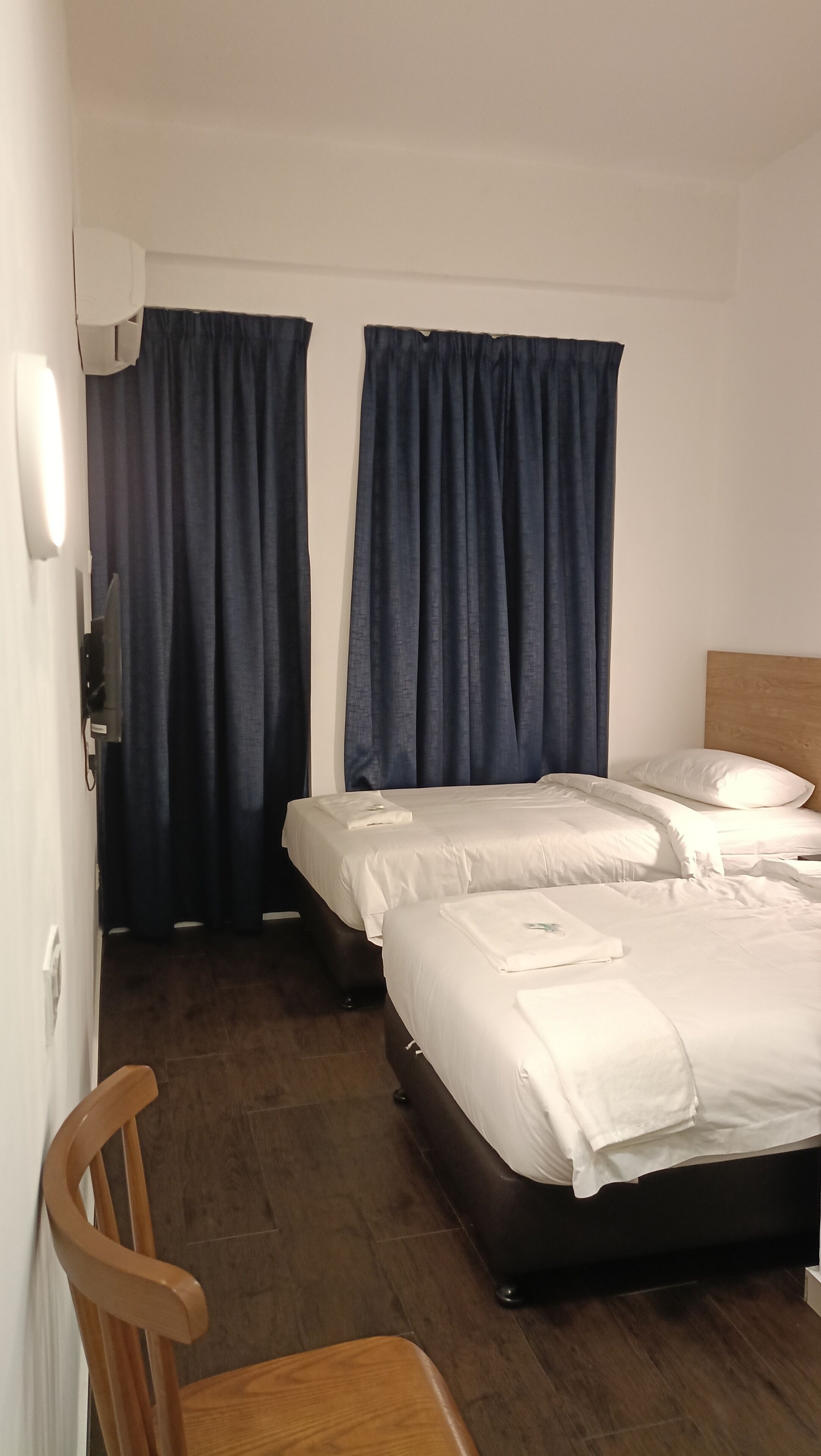 Habitación de hotel con dos camas, cortinas oscuras, cabecera de madera, una silla de madera y un televisor de pantalla plana.