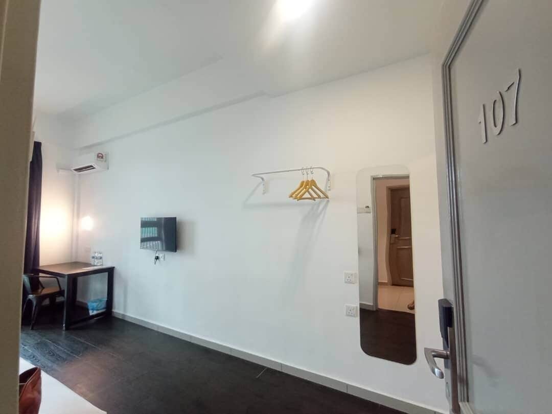 Habitación de hotel con piso oscuro, paredes blancas, televisor de pantalla plana montado en la pared, una mesita con una silla, un perchero con colgadores y una puerta con el número 107.
