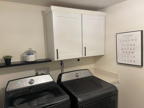 A laundry room with two front-loading washers, a white cabinet above, a shelf with a plant and detergent, and a laundry symbols chart on the wall.