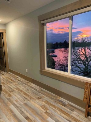 A room with a large window overlooking a body of water and trees during sunset.