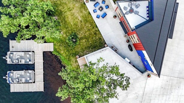 An aerial view of a property with a dock, two boats, a patio with outdoor furniture, and a building with a red and blue roof.