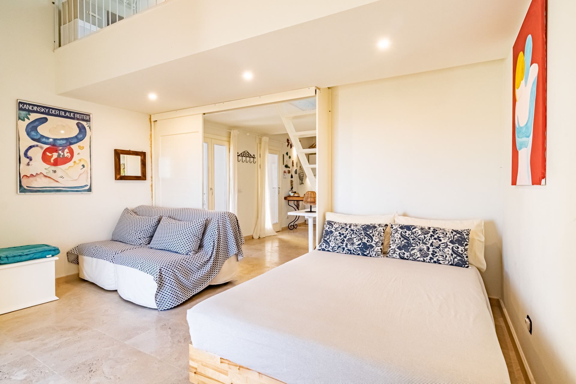 A spacious bedroom with a large bed, a sofa, and a small table. There is a staircase leading to an upper level.