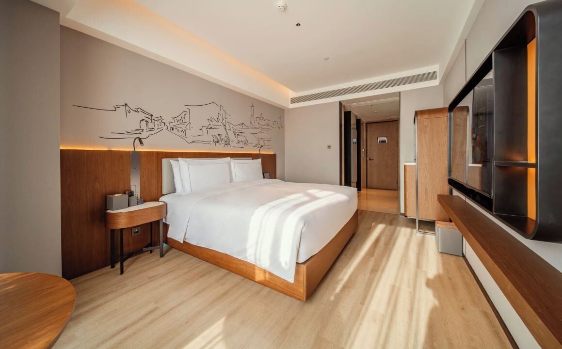 A modern hotel room with a large bed, wooden headboard, bedside table, and a flat-screen TV.