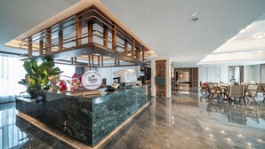 Un hall d’hôtel moderne avec un comptoir d’accueil en marbre, une grande plante en pot et un espace détente avec des chaises et des tables en bois.