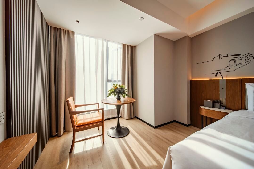 A hotel room with a bed, a desk, a chair, a round table with a plant, and a window with curtains.