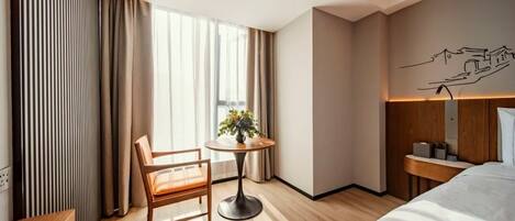 A hotel room with a bed, a desk, a chair, a round table with a plant, and a window with curtains.