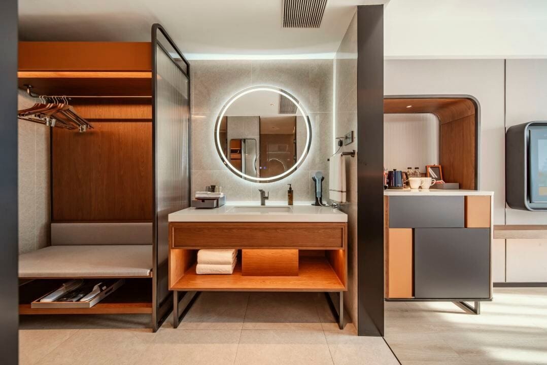 A modern bathroom with a wooden vanity, a round mirror with lighting, a built-in shower, and a separate vanity with a sink and mirror.