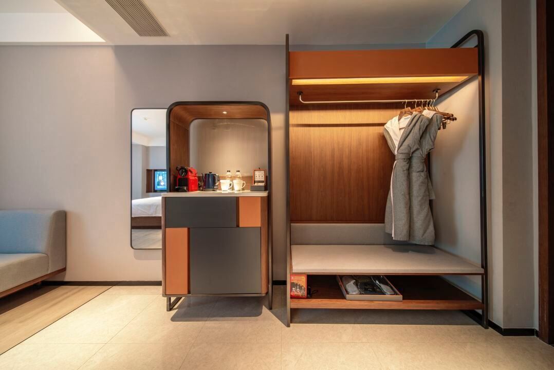 Een moderne hotelkamer met een ingebouwd bureau, een spiegel, een garderobe met kledingroede en een bankje.