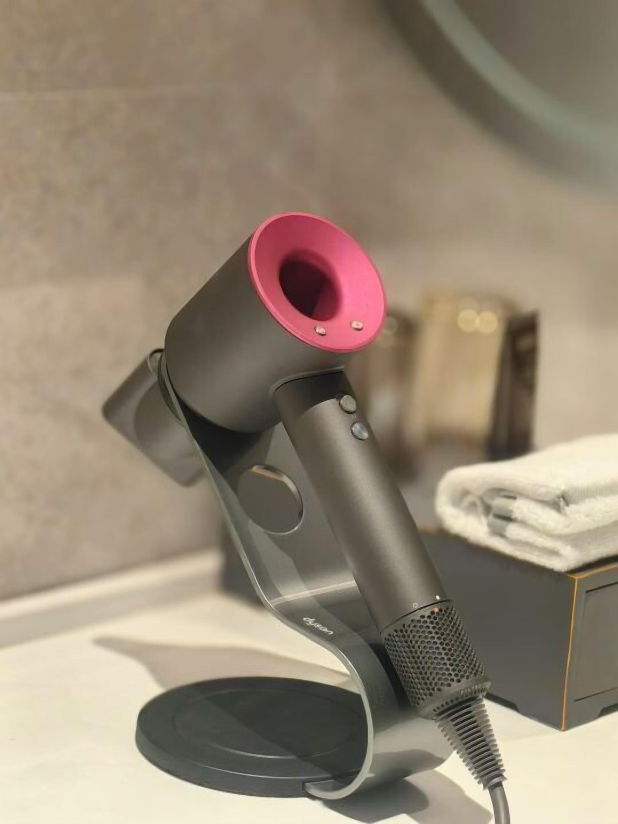 A hair dryer with a pink nozzle and a black cord, placed on a white surface with a towel and a box in the background.