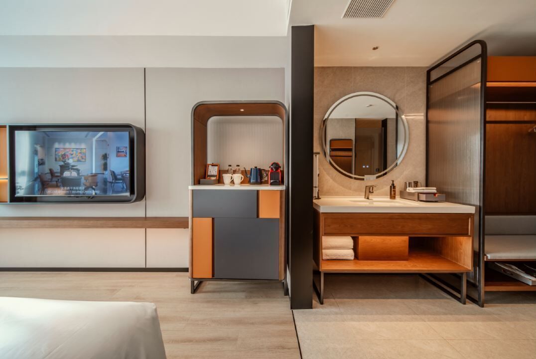 A modern hotel room with a large bed, a TV mounted on the wall, a wooden vanity with a mirror, and a wooden cabinet.