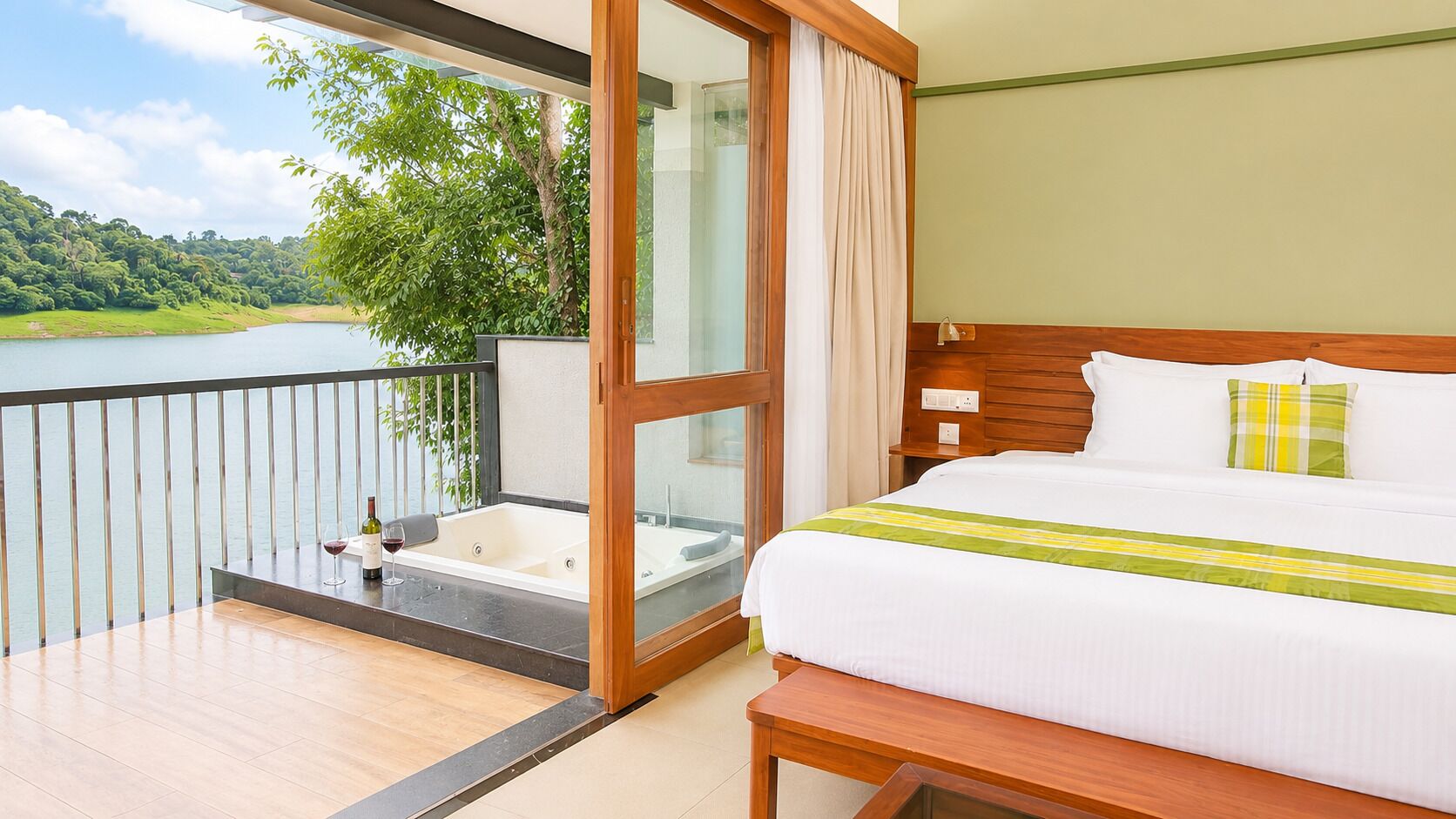 A hotel room with a balcony, a bed, a bathtub, and a view of a lake and greenery.