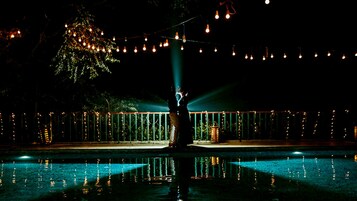 Escena nocturna al aire libre con piscina, luces colgantes y un árbol.