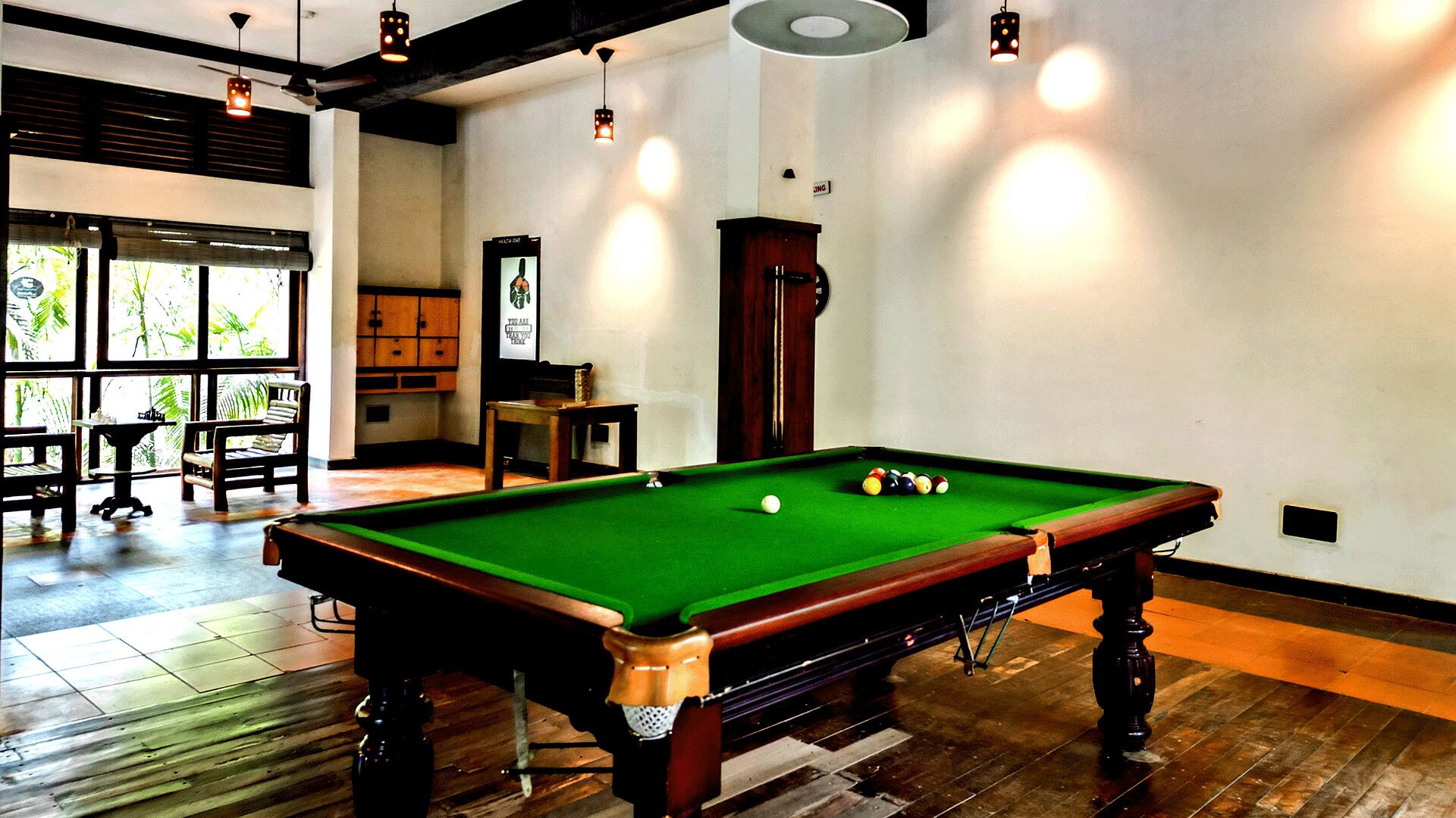 A pool table in a room with wooden flooring, a ceiling fan, and a large window with a view of greenery outside.